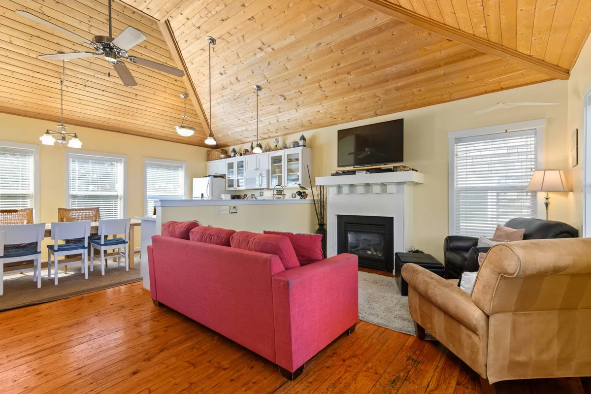 A warm, inviting open-concept space featuring a pink sofa in the living area, a visible dining area with ample seating, a cozy gas fireplace, and a modern kitchen counter in the background. The scene is bright, with a family-oriented layout that visually connects all three major spaces for gathering and relaxation.