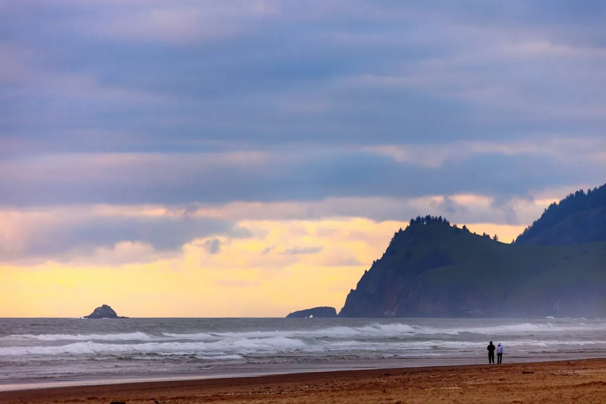 The image captures a breathtaking view of the Oregon Coast at sunset, with waves rolling in, distant cliffs, and a colorful sky. Silhouetted figures stroll along the sandy beach, encapsulating the area’s natural beauty and the sense of exploration.