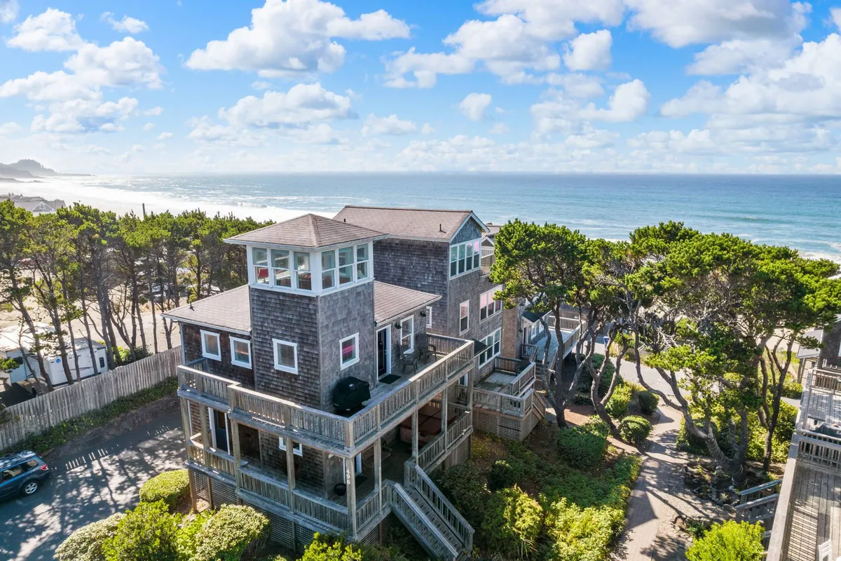 The image showcases Luna Bella, a charming two-story coastal home set among mature trees, with a spacious deck overlooking the Pacific Ocean. The house’s distinctive windowed tower is visible, hinting at panoramic views, while inviting outdoor furniture on the deck suggests relaxation. The scene captures the soft glow of sunset, with a welcoming path leading toward the beach.