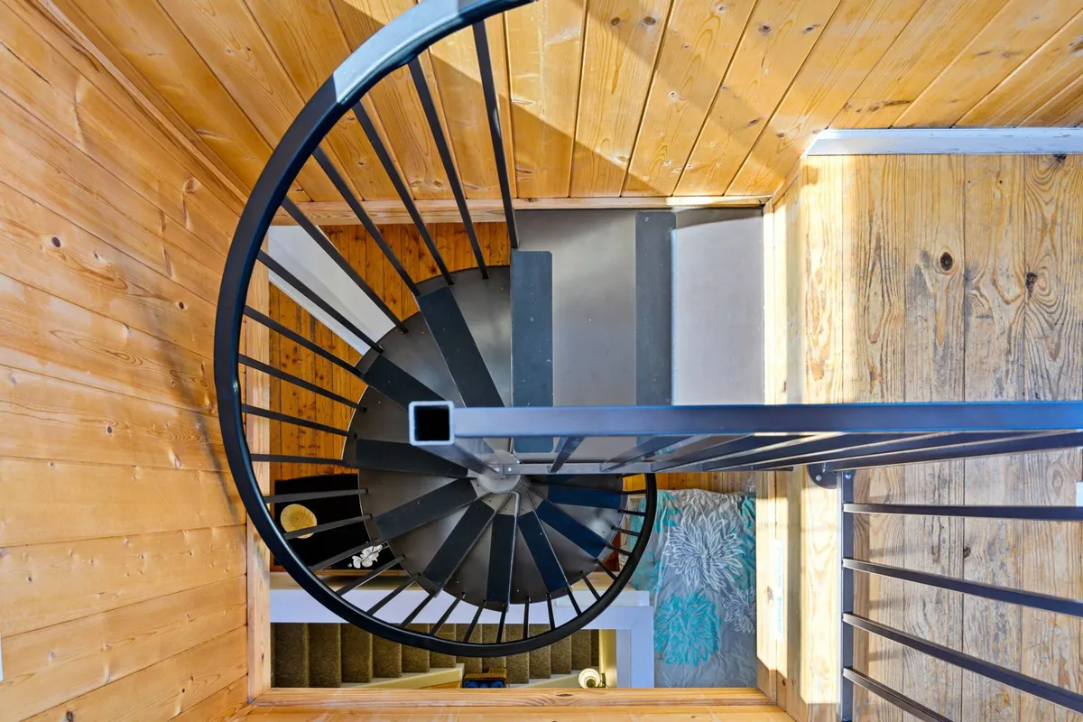 A close-up interior shot of the spiral staircase, with warm wooden walls and contemporary textured flooring. The perspective gives a sense of ascent, emphasizing the journey and anticipation of what lies above.