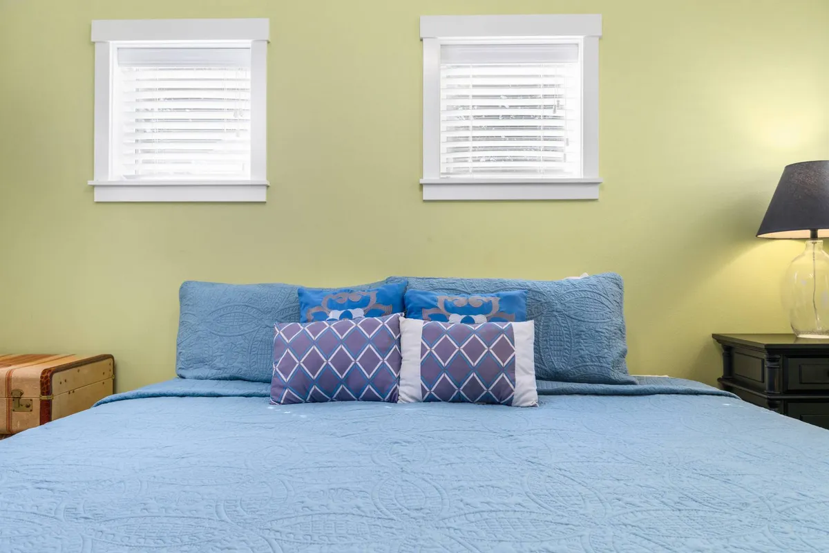 A cozy bedroom with a large bed dressed in blue bedding and decorative pillows, complemented by a nightstand with a lamp and multiple windows letting in natural light, evoking a serene and restful ambiance.
