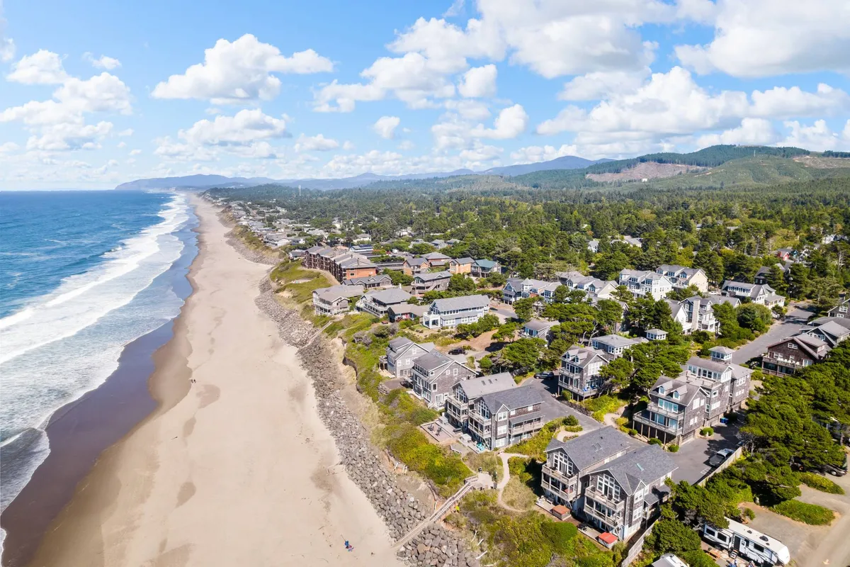 A sweeping view of a wide sandy beach with gentle ocean waves, coastal homes dotting the shoreline, lush greenery, and distant mountains under a relaxed, inviting sky. The scene captures the essence of a family-friendly beach, ideal for sandcastle building, surfing, and exploring.