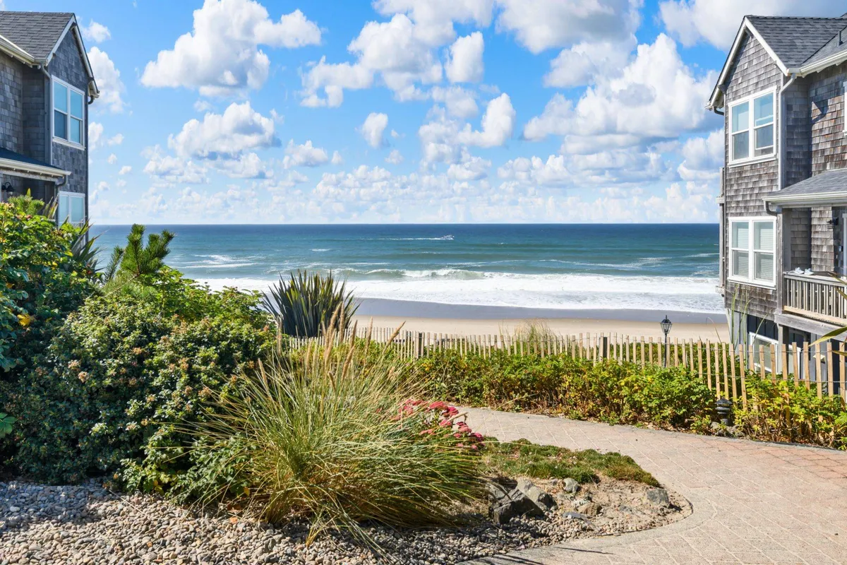 A tranquil garden with lush landscaping opening onto a direct path to the sandy beach and ocean, beneath a cloudy sky. The scene suggests a peaceful transition from home life to beach adventures, with opportunities for beach walks and nature appreciation.