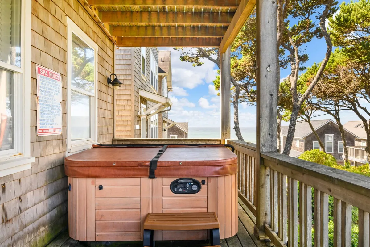 This image features an outdoor hot tub situated on a deck, surrounded by lush greenery, with a stunning view of the sea visible beyond the railing. The tranquil setting is enhanced by the warm lighting, inviting guests to unwind while enjoying the ocean vista and the sound of waves.