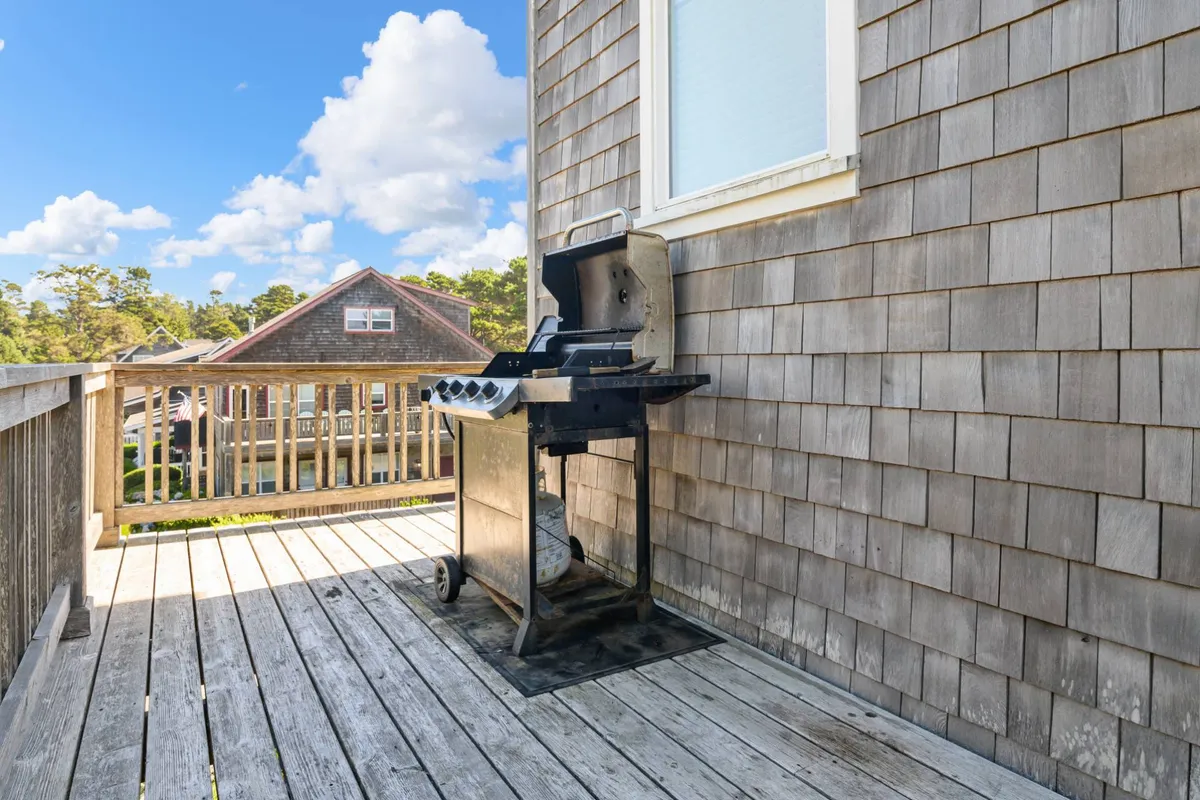 This image features a wooden deck with a grill surrounded by lush greenery, highlighting the space for outdoor dining and relaxation. The inviting setup captures the ambiance of al fresco meals and sunset gatherings, pivotal to the section's mention of the wrap-around porch and BBQ area.