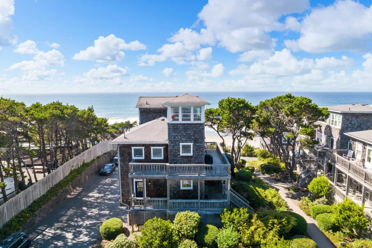 A broad exterior view of the house, featuring the wrap-around deck and direct sightlines to the ocean and landscaped surroundings. The scene is bathed in soft light, evoking a serene, welcoming mood and illustrating the deck's connection to the neighborhood and natural beauty.